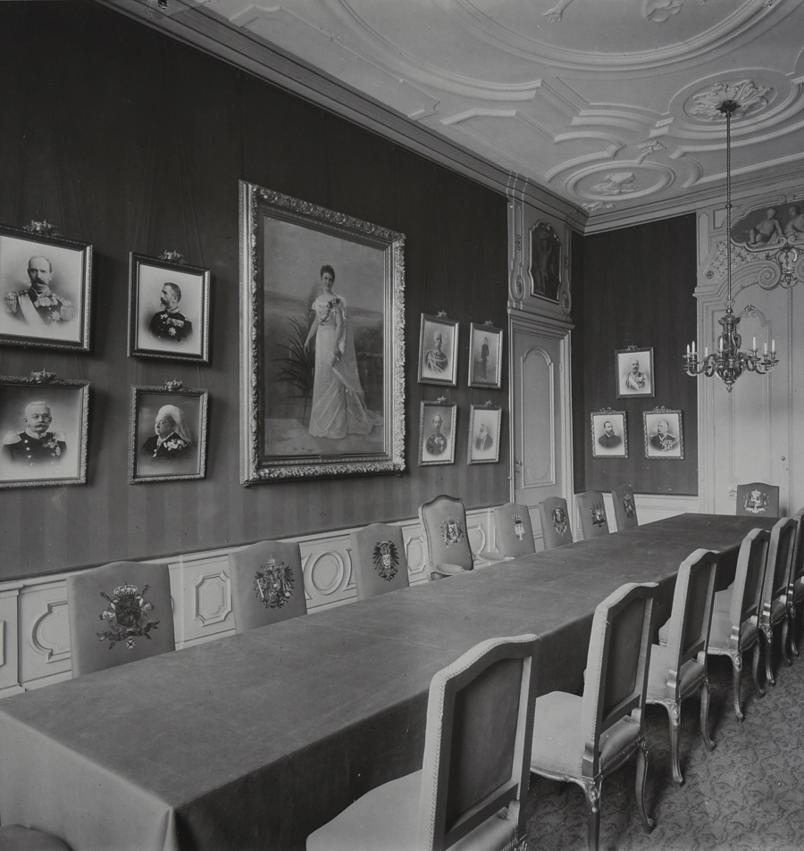 Interieurfoto van de zittingzaal van het Permanent Hof van Arbitrage met zicht op de tussendeuren en de wand met daarop een groot portret van koningin Wilhelmina. Links, op de onderste rij, zijn de portretten van Victoria, koningin van Groot-Brittannië en van Adolf, groothertog van Luxemburg herkenbaar. Foto uit een portefeuille met een serie van zes zwart-wit foto's van het Permanent Hof van Arbitrage dat van 1901 tot de opening van het Vredespaleis in 1913 was gevestigd aan de Prinsegracht 71 in Den Haag.